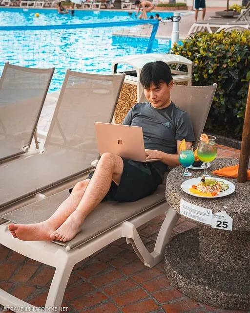 Travelling Foodie Raymond Cua lying on the pool chair using Microsoft Surface Laptop 3 with cocktails