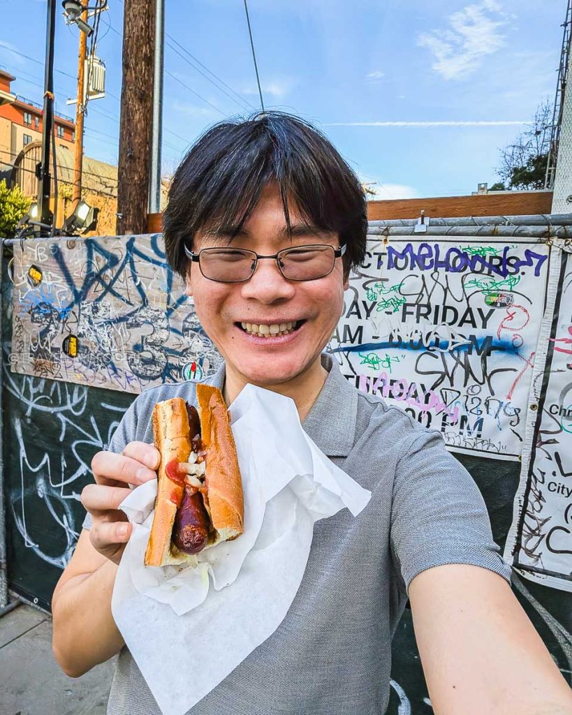 Travelling Foodie Raymond Cua enjoying an iconic sausage outside Top Dog, a historic street food spot and highly rated place to eat in Berkeley.