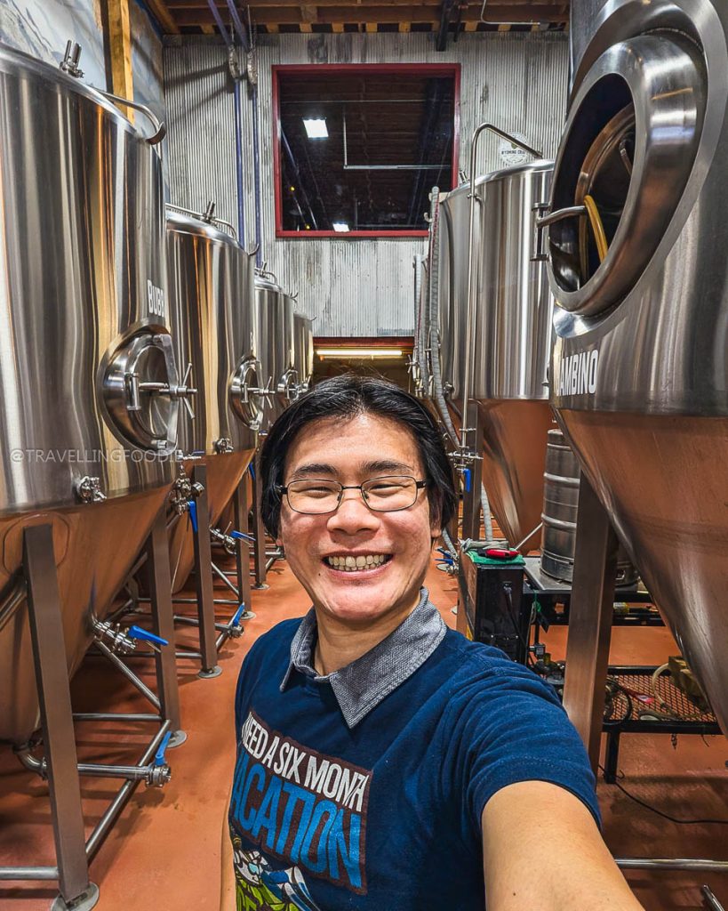 Travelling Foodie Raymond Cua smiling next to massive beer fermentation tanks at Bond's Brewing Company in Laramie Wyoming.
