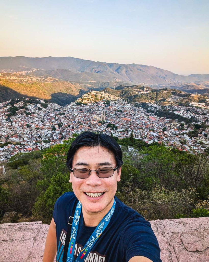 Raymond Cua smiling with a stunning panoramic view of the Pueblo Magico town of Taxco de Alarcon from above.