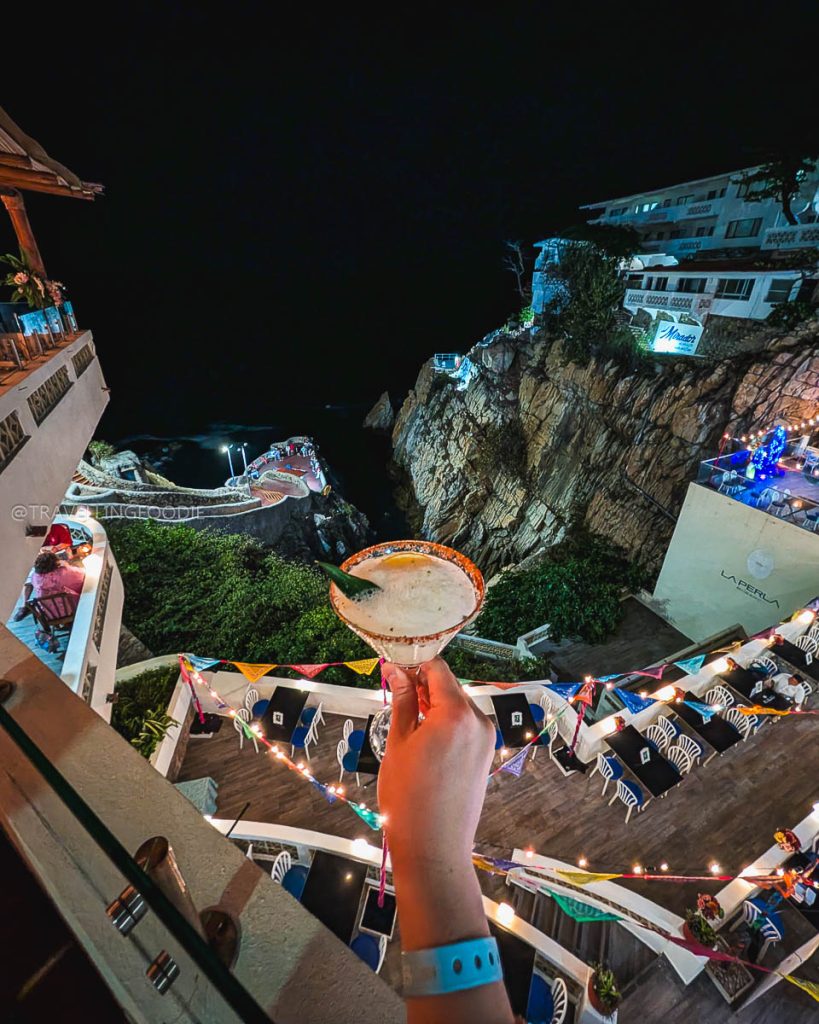 Hand holding a cocktail at Hotel Mirador with a stunning night view of La Quebrada cliff diving spot in Acapulco.
