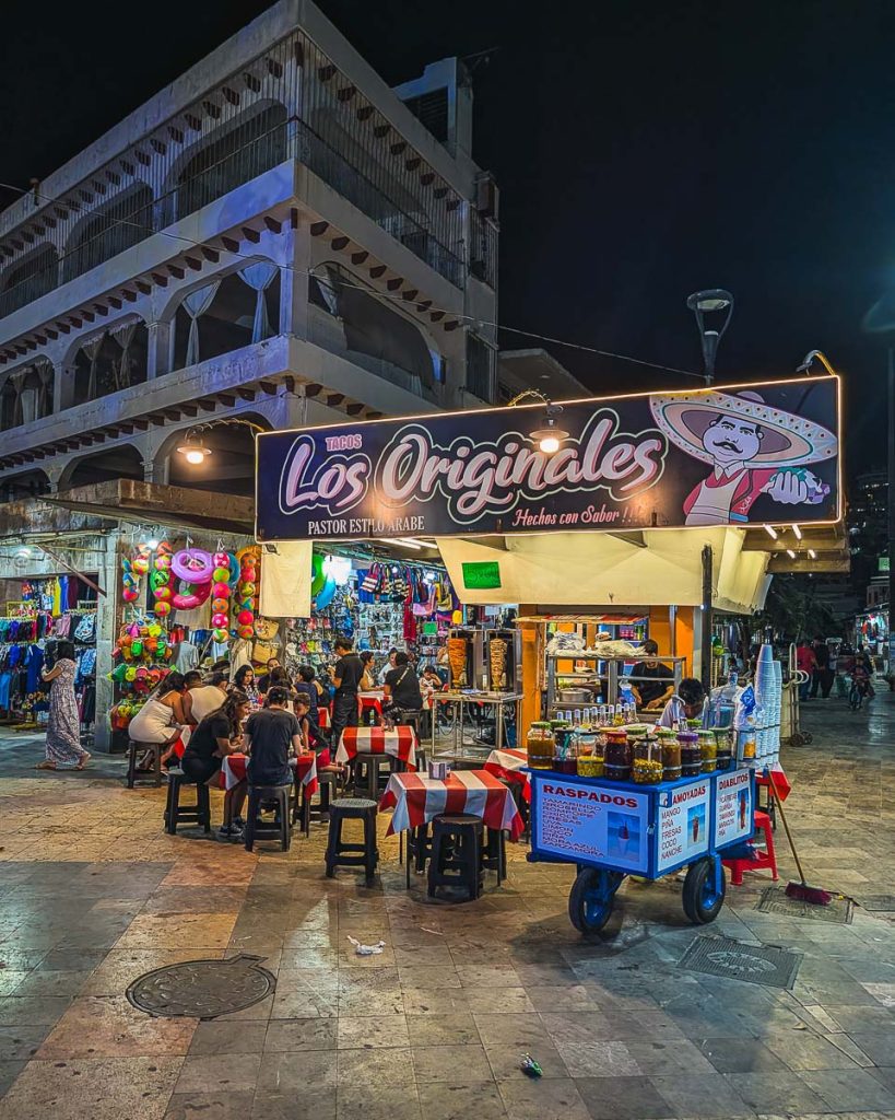 Lively night scene at Plaza Alvarez featuring a colorful Tacos Los Originales stand and a raspados cart in Acapulco.
