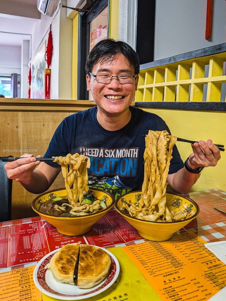 Travelling Foodie Raymond Cua holding wide hand-ripped noodles at Number 1 Noodle House to show where locals eat in Saskatoon.