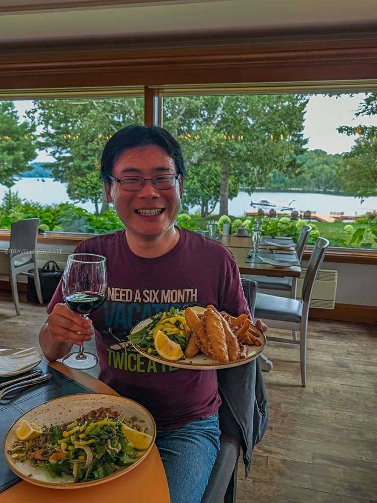 Travelling Foodie Raymond Cua holding fish and chips with wine at Auberge du Lac-a-l'Eau-Claire during a Mauricie culinary road trip.