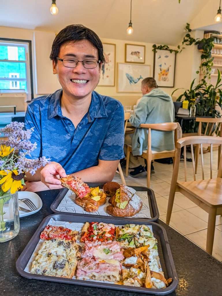Raymond Cua holding a slice of Roman pizza al taglio beside pastries at Roberto Pizza Romana in Chelsea Quebec.